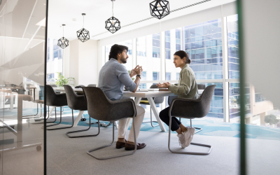 Two individuals sit at a table in a modern office with large glass windows. One informs the other about being impacted by layoffs and the offer of outplacement services. The impacted individual asks, "What are outplacement services?" 