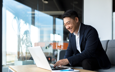 A smiling leader in a business suit works at his desk with an open laptop, showcasing his leadership skills identified through his company’s leadership assessments.