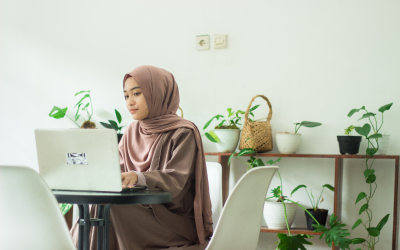 A woman sits at her dining room table with a laptop, surrounded by house plants, working on building a personal brand on LinkedIn. 