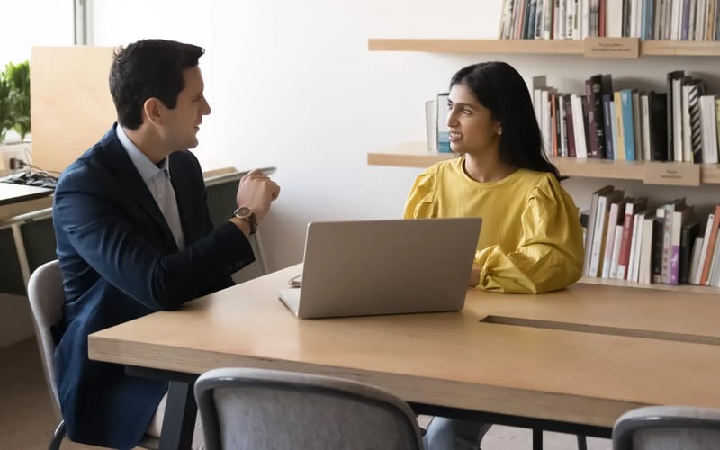 A manager explains something to a woman in yellow shirt. They are in an office with a wall of books in background.