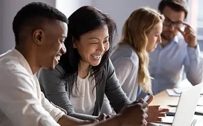 Employees collaborate and smile around a laptop, reflecting the impact of effective engagement strategies in the workplace.