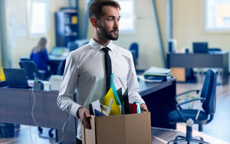 Man in shirt and tie in need of career transition support. He is in an office setting & carries a box of office supplies. 