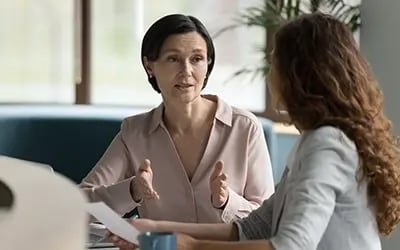 Leadership development session featuring a white woman in tan blouse explaining something to younger woman who faces away from camera.
