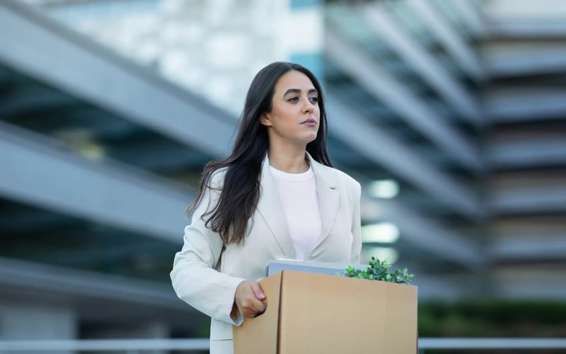 A woman going through a career transition walks away from an office building while carrying a box of her workplace items. 