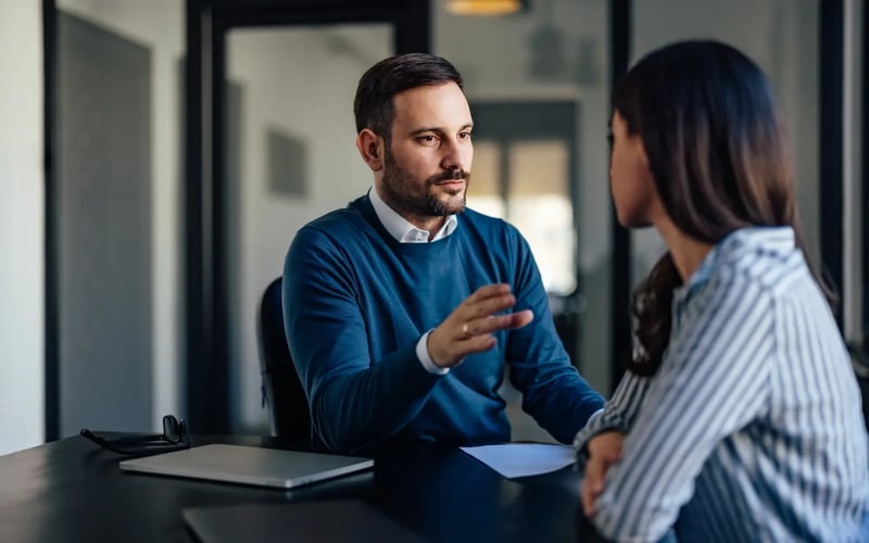 A white man with trimmed beard and a blue sweater is explaining something to a woman in striped shirt whose back is to the camera. 
