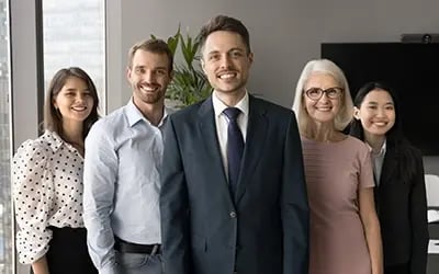 Inclusive leadership featuring a mixed-race man in business suit flanked by older woman, Asian woman and two white people in office setting. 