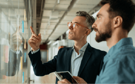 Two business professionals, a leadership coach and a colleague, examining a glass wall in a contemporary office.