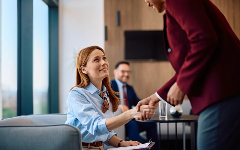 A mid-30s white woman in blue blouse is seated in a reception area. A man in burgundy blazer and blue pants is bending down to shake hands. Both are smiling.